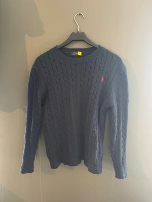 Mörkblå kabelstickad tröja Polo Ralph Lauren - Kabelstickad mörkblå tröja från Polo Ralph Lauren med rund halsringning och den klassiska röda logotypen broderad på bröstet. Tröjan har långa ärmar och ribbade muddar vid ärmslut och nederkant. Perfekt för lager på lager under kyliga dagar. Hör av er vid frågor och priser✅👍