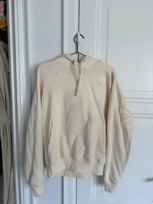 Nude projects - vit hoodie med ficka och huva - Säljer en vit hoodie med stor magficka och huva. Hoodien har ribbade muddar vid ärmar och midja samt en unik detalj på ena ärmen. Perfekt för en avslappnad och stilren look. Tryck på ryggen