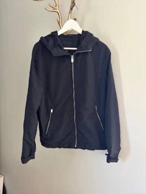 Svart vindjacka med huva - Snygg svart vindjacka med dragkedja framtill och två fickor med dragkedja. Jackan har en klassisk huva och elastiska muddar vid ärmsluten. Tillverkad i lätt och vindtätt material, perfekt för blåsiga dagar.