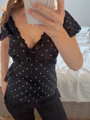 Svart prickig blus med spets och snörning - Snygg svart blus med vita prickar, djup v-ringning och spetsdetaljer längs urringningen. Blusen har korta ärmar, snörning framtill och en feminin passform. Perfekt för dig som gillar trendiga detaljer och en söt look.