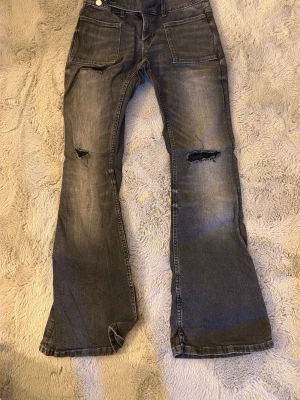 Grå slitna bootcut jeans - Ett par grå bootcut jeans med slitningar och hål på knäna. Jeansen har stora fickor fram och bak, samt en lätt tvättad look för en avslappnad stil.