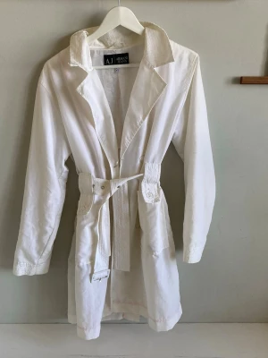 Vit trenchcoat från Armani Jeans - Stilren vit trenchcoat från Armani Jeans med klassisk krage, knytskärp i midjan och dekorativa knappar vid fickorna. Lätt material som ger en avslappnad men ändå elegant look. Perfekt för dig som vill ha en tidlös och fräsch jacka.