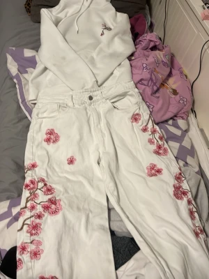 Vita jeans med rosa blommor - Unika vita jeans med broderade rosa blommor och grenar längs benen. Rak passform och klassisk femficksmodell. Perfekta för dig som vill sticka ut med en kreativ och trendig look.