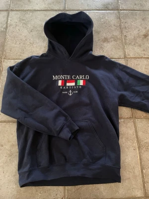 Mörkblå hoodie Monte Carlo Kartisto - Mörkblå hoodie från Kartisto med broderad text 'Monte Carlo' och flaggor på bröstet. Klassisk känguruficka framtill och stor huva. Perfekt för en chill och sportig stil. Materialet känns mjukt och bekvämt.