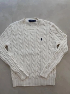 Ralph lauren stickad vit  - Kabelstickad vit tröja från Polo Ralph Lauren med rund halsringning och lång ärm. Tröjan har det klassiska broderade logotypen i marinblått på bröstet och ribbade muddar vid ärmslut och nederkant. Perfekt för en clean och stilren look.