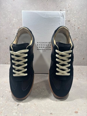 Svarta sneakers Maison Margiela - Stilrena svarta sneakers från Maison Margiela med beigea skosnören och gummisula. Ovandelen är i mocka och läder, med klassisk låg silhuett och diskreta detaljer. Perfekta för dig som gillar minimalistisk och trendig design.
