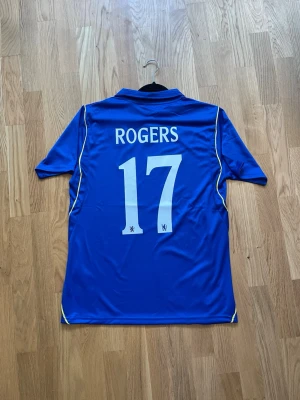 Rogers #17 - M - Säljer en blå Chelsea matchtröja från Nike med vita detaljer och nummer 17 samt namnet ROGERS på ryggen. Tröjan har korta ärmar, krage och klubbmärke på bröstet. Perfekt för fotbollsträning eller att bära på läktaren.