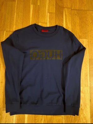 Mörkblå sweatshirt från HUGO - Snygg mörkblå sweatshirt från HUGO med stor logga i svart på bröstet. Tröjan har rund halsringning och ribbade muddar vid ärmslut och nederkant. Perfekt för en avslappnad och trendig stil.