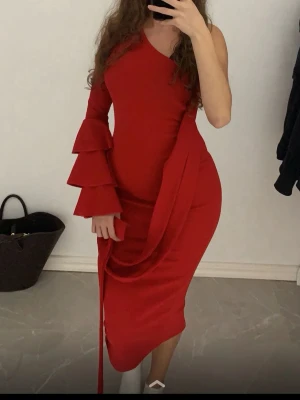Röd oneshoulder klänning med volangärm - Säljer en röd oneshoulder klänning med asymmetrisk skärning och långa volangärmar. Klänningen har en figurnära passform och en snygg detalj med tyg som hänger ner från midjan. 