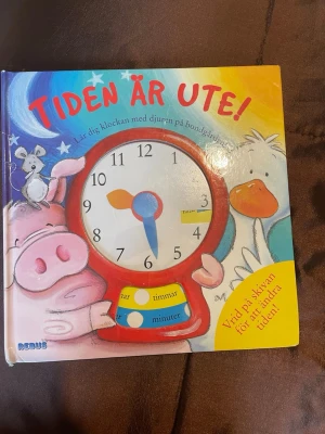 Tiden är ute! interaktiv barnbok - En färgglad och interaktiv barnbok med titeln 'Tiden är ute!' där du lär dig klockan med djuren på bondgården. Boken har en inbyggd urtavla med rörliga visare och tydliga illustrationer av djur. Perfekt för att öva på att läsa av tiden på ett lekfullt sätt.