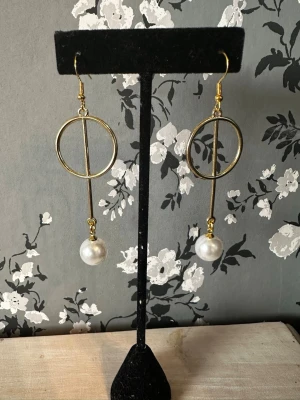 Guldörhängen med pärldetalj - Säljer ett par trendiga örhängen i guldfärgad metall med geometrisk design. Örhängena har en cirkel och en rak stav, samt en vit pärla som hänger längst ner. Perfekta för dig som gillar modern och stilren look.