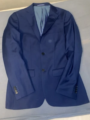 Dressman Mörkblå kostym med kavaj och byxor - Snygg mörkblå kostym med klassisk kavaj och matchande byxor. Kavajen har två knappar framtill, fickor och knappdetaljer vid ärmsluten. Perfekt för dig som vill ha en stilren och tidlös look. Från dressman 