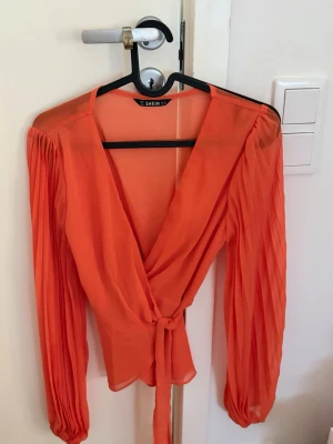 Orange omlottblus  - Trendig orange omlottblus från SHEIN med långa, plisserade ärmar och knytband i midjan. Blusen är gjord i ett tunt, transparent material som ger en luftig känsla och har en djup v-ringning framtill.