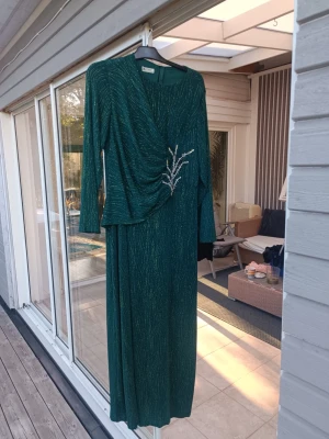 Festklänning - Elegant grön långklänning med glittriga inslag och omlott-detalj framtill. Klänningen har långa ärmar och en dekorativ broderad detalj med pärlor på sidan. Perfekt för dig som vill sticka ut med stil. Storlek 48/50