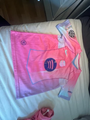 Rosa Nike fotbollströja Raphinha 11 - Snygg fotbollströja i rosa och lila toner från Nike med tryck av Raphinha och nummer 11 på ryggen. Tröjan har korta ärmar, flera loggor och klubbmärke framtill samt detaljer i vitt och blått. Perfekt för dig som älskar fotboll och vill sticka ut på planen.