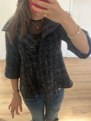 Gråmelerad bouclé kappa med 3/4-ärm - Säljer en gråmelerad bouclé kappa med 3/4-ärm och bred krage. Kappan har två svarta fickor framtill och stängs med dragkedja. Materialet är tjockt och strukturerat, perfekt för kyligare dagar. Snygg och trendig modell som passar till jeans.