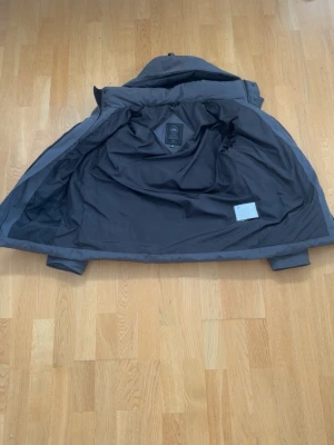 Svart Canada Goose black label 🇨🇦 ”äkta” Oanvänd  - Snygg mörkblå dunjacka med avtagbar huva och quiltad design. Jackan har en patch på ärmen och stängs med både dragkedja och knappar. Perfekt för kalla vinterdagar tack vare det tjocka fodret. Klassisk och stilren modell som håller dig varm.