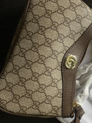 Beige och brun axelväska från Gucci - Snygg axelväska från Gucci i beige canvas med klassiskt GG-mönster och bruna läderdetaljer. Väskan har guldfärgad logga framtill och dragkedja. Perfekt för dig som gillar tidlös design och lyxiga detaljer.