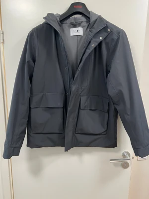Mörkgrå regnjacka från NN07 - Snygg mörkgrå regnjacka från NN07 med huva och stora framfickor. Jackan är tillverkad i stretchigt polyestertyg med PU-laminering, har tejpade sömmar och är både vind- och vattentät. Fodrad med fukttransporterande material och Primaloft Gold vaddering. Jackan har använd ett par gånger bara.