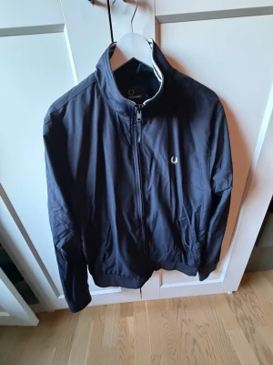 Svart vindjacka från Fred Perry - Snygg svart vindjacka från Fred Perry med klassisk logga broderad på bröstet. Jackan har dragkedja framtill, hög krage och ribbade muddar vid ärmslut och nederkant. Perfekt för dig som vill ha en stilren och enkel jacka.