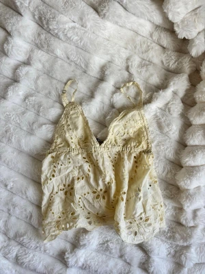 Gul linne med broderi - Säljer en söt beige linnetopp med tunna axelband och v-ringning. Toppen har fina broderade detaljer och spets längs kanterna, vilket ger en romantisk känsla. Perfekt att styla med jeans eller kjol för en trendig look.