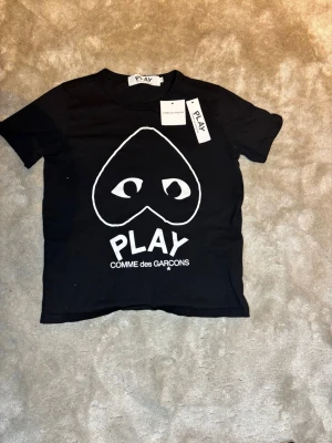 Svart Comme des Garçons PLAY t-shirt - Svart t-shirt från Comme des Garçons PLAY med det ikoniska hjärtmotivet och ögon i vitt framtill. Klassisk rund hals och korta ärmar. Perfekt för dig som gillar streetwear och vill sticka ut med en stilren men ändå lekfull design. Storlek S men passar som XS