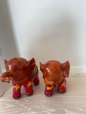 Dekorativa elefanter i trä - Två handgjorda elefanter i trä med röd och gul dekor. Elefanterna har fina detaljer, blommönster på benen och en av dem har ett dekorerat sadelparti. Perfekt som inredningsdetalj för dig som gillar unika prydnader.
