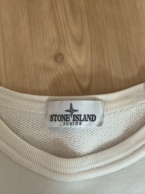Vit tröja från Stone Island Junior - Vit tröja från Stone Island Junior med klassisk rund hals och ikonisk logotyp-patch på ärmen. Tillverkad i mjuk bomull som känns skön mot huden. Perfekt för en clean och stilren look.