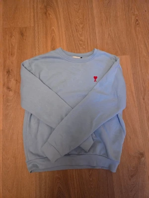 Ljusblå Ami Paris sweatshirt - Ljusblå sweatshirt med rund hals och ett litet rött hjärtemärke broderat på bröstet. Tröjan har långa ärmar och ribbade muddar vid ärmslut och nederkant. Perfekt för en avslappnad och stilren look.