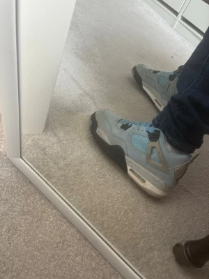 Jordan 4 retro unc skor - Snygga sneakers i ljusblått med svarta och grå detaljer samt vit sula. Skorna har meshpaneler och synliga snörhål i plast. Tjock sula med luftbubbla och cool design som sticker ut. Perfekta för dig som gillar streetwear och vill ha något unikt på fötterna. Perfekta skor nu när hösten och vintern kommer