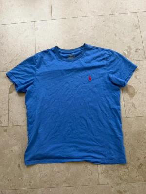 Blå t-shirt från Polo Ralph Lauren - Snygg blå t-shirt från Polo Ralph Lauren med klassisk röd broderad logga på bröstet. T-shirten har rund halsringning och korta ärmar. Tillverkad i mjuk bomull som känns skön mot huden. Perfekt för en avslappnad och stilren look.