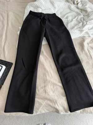 Svarta vida mjukisbyxor - Säljer ett par svarta mjukisbyxor med raka, vida ben och resår i midjan med dragsko. Perfekta för chill dagar eller när du vill ha det extra bekvämt. Tillverkade i mjukt material som känns skönt mot huden.