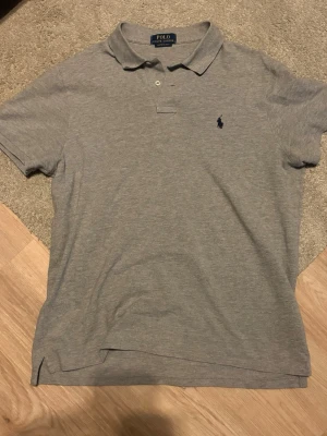 Grå pikétröja från Polo Ralph Lauren - Klassisk grå pikétröja från Polo Ralph Lauren med korta ärmar och krage. Tröjan har två knappar vid halsen och den ikoniska broderade loggan i marinblått på bröstet. Tillverkad i mjuk bomull för en bekväm känsla.