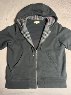 Burberry - Snygg mörkgrå hoodie från Burberry med dragkedja, klassiskt rutigt foder i huvan och broderad logga på bröstet. Två stora fickor framtill och justerbar huva. Perfekt för dig som gillar stilrena detaljer och premiumkänsla.