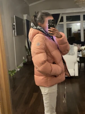 Rosa dunjacka från Moncler - Säljer en fluffig rosa dunjacka från Moncler med stor huva och svart logotypdetalj på kragen. Jackan har dragkedja framtill, två sidofickor med svarta dragkedjor och en klassisk Moncler-logga på ärmen. Perfekt för kalla vinterdagar.