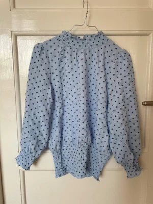 Ljusblå blus från Neo Noir - Supersöt ljusblå blus med små svarta blommor och hög krage. Blusen har långärmade puffärmar med smock vid ärmslut och midja. Perfekt för dig som gillar romantiska detaljer och en feminin look.