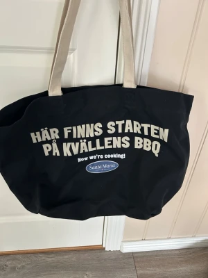 Svart tygkasse med tryck Santa Maria - Stor svart tygkasse med beige handtag och trycket 'Här finns starten på kvällens BBQ' samt Santa Maria-logga. Perfekt för att bära med sig grejer till grillkvällen eller stranden. Rymlig och enkel att slänga över axeln. Helt ny