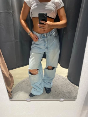 Ljusa baggy jeans med hål på knäna - Säljer ett par ljusblå baggy jeans med stora slitningar över båda knäna. Jeansen har hög midja, raka ben och klassisk femficksdesign. Perfekta för en avslappnad och trendig streetstyle-look.