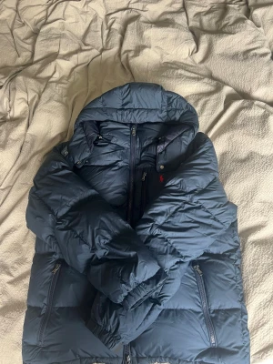 Mörkblå pufferjacka från Polo Ralph Lauren - Snygg mörkblå pufferjacka från Polo Ralph Lauren med röd broderad logga på bröstet. Jackan har dragkedja framtill, två sidofickor med dragkedja och en hög krage med tryckknappar. Perfekt för kalla vinterdagar och riktigt varm.