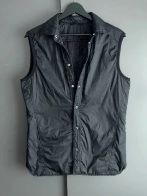 Svart dunväst med tryckknappar - Stilren svart dunväst med hög krage och tryckknappar framtill. Västen har en klassisk passform och är tillverkad i ett glansigt material som ger en modern look. Perfekt att slänga över en hoodie eller tröja för extra värme under kyliga dagar.