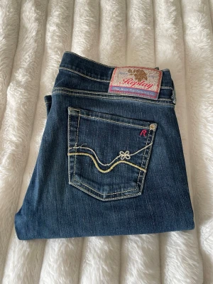 Blå jeans från Replay med brodyr - Säljer ett par klassiska blå jeans från Replay med snygga kontrastsömmar och broderad bakficka. Jeansen har normal midja, raka ben och detaljer i gult och lila. Materialet är slitstark denim i bomull. Perfekta för en trendig och avslappnad look.