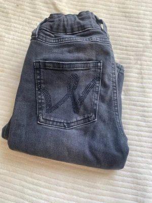 Grå jeans med strassdetalj Gina Tricot - Snygga grå jeans från Gina Tricot med strassdetalj i form av ett W på bakfickan. Jeansen har hög midja, klassisk femficksmodell och bälteshällor. Materialet är stretchigt denim för extra komfort och passform.