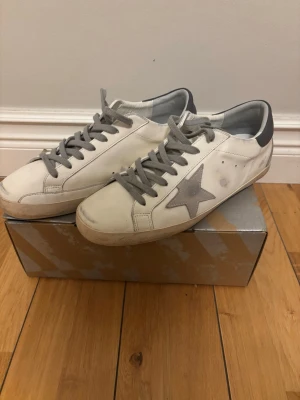 Golden Goose vita sneakers med grå stjärna - Golden Goose sneakers i vitt läder med grå mockadetaljer, grå stjärna på sidan och grå snörning. Hälpartiet är mörkgrått med logotyp och baksidan har mocka. Klassisk låg modell med slitna detaljer för en trendig look.