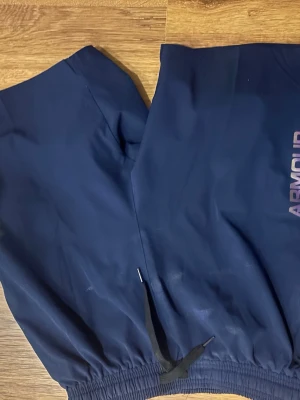 Marinblå shorts Under Armour - Mörkblå mjukisbyxor från Under Armour med resår i midjan och svarta dragsnören. Byxorna har logga på benet och är tillverkade i ett mjukt, sportigt material som passar perfekt för chill eller träning.