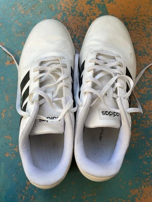 Vita Adidas sneakers med svarta ränder - Klassiska vita sneakers från Adidas med de ikoniska tre svarta ränderna på sidan. Skorna har lågt skaft, vit sula och snörning framtill. Tillverkade i syntetmaterial och textil för en clean och sportig look.