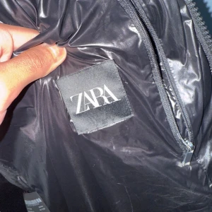 Svart pufferjacka från Zara - Säljer en svart pufferjacka från Zara med glansig finish och huva. Jackan har dragkedja framtill och två sidofickor. Perfekt för kalla dagar när du vill hålla stilen och samtidigt vara varm.