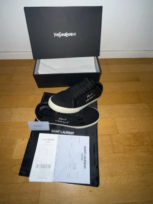Svarta Saint Laurent sneakers - Svarta sneakers från Saint Laurent med vit sula och handskriven logga på sidan. Klassisk låg modell med snörning och canvasmaterial. Kommer med originalkartong, dustbag och äkthetsintyg. Perfekta för dig som gillar stilrena och exklusiva skor.