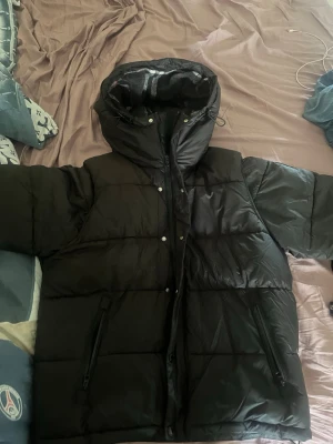 Svart pufferjacka från Burberry - Snygg svart pufferjacka från Burberry med huva och dragkedja framtill. Jackan har två stora fickor med dragkedja och Burberry-logga på ärmen. Insidan av huvan har klassiskt rutmönster. Perfekt för kalla vinterdagar. passar L beroende på L och vikt 