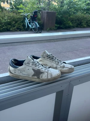 Golden Goose vita sneakers med stjärna - Golden Goose sneakers i vitt läder med grå mockadetaljer och en ikonisk glittrig stjärna på sidan. Skorna har mörkblå hälkappa och klassisk låg siluett med snörning. Perfekta för en avslappnad streetstyle-look.