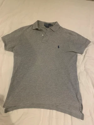 Grå pikétröja från Polo Ralph Lauren - Säljer en klassisk grå pikétröja från Polo Ralph Lauren med blå logga broderad på bröstet. Tröjan har korta ärmar, krage och knappar vid halsen. Tillverkad i mjuk bomull och har en normal passform. Perfekt till en avslappnad stil.